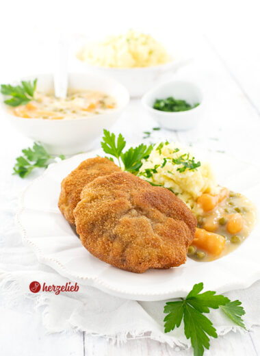 Zwei panierte Hackfleischschnitzel auf einen weißen Teller. Angerichtet mit Kartoffelbrei und Erbsen und Wurzeln in weißer Soße, dekoriert mit Petersilie. Im Hintergrund eine Schüssel mit Kartoffelmus, eine mit Gemüse in weißer Sauce und ein Schälchen mit gehackter Petersilie