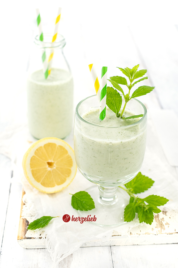 Ein Glas mit Melonen-Lassi im Vordergrund. Dekoriert mit Zitronenmelisse und zwei Trinkhalmen. Im Hintergrund steht eine Flasche mit dem Joghurtdrink, in dem ebenfalls zwei Trinkhalme stecken. Davor liegt eine halbe Zitrone.