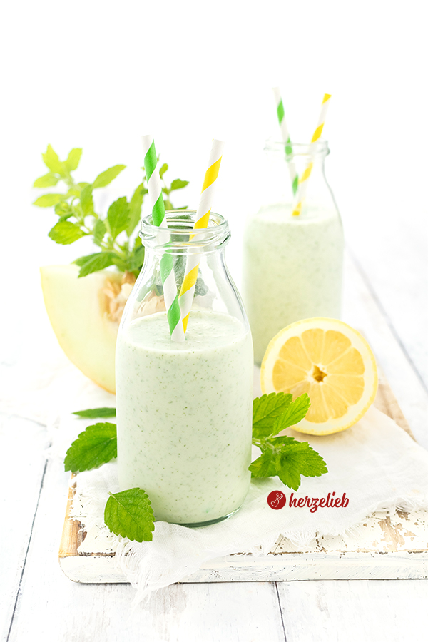 Zwei kleine Flachen mit Melonen-Lassi, dekoriert jeweils mit Zitronenmelisse und zwei Trinkhalmen. Im Hintergrund ein Melonenstück, eine Zitrone und Zitronenmelissezweige. 