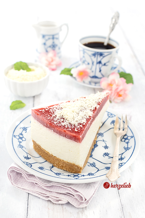 Ein Stück Quark-Mascarpone-Torte auf einem blauweißen Teller. Auf dem Tellerrand liegt eine Kuchengabel. Die Torte ganz oben eine Schicht aus Erdbeermus und ist mit Raspeln aus weißer Schokolade. Im Hintergrund Kaffeegeschirr, eine Schale mit geraspelter, weißer Schokolade und ein Milchkännchen sowie rosa Blüten.