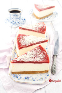 Drei Stücke Quark-Mascarpone-Torte auf einer einer weißen Porzellanplatte mit blauen Blümchen. Jedes Stück hat oben auf Erdbeermus und ist mit weißen Schokorapseln bestreut. Rechts neben dem Teller liegt ein Messer. Im Hintergrund ein Kuchenstück auf einem Teller und eine Tasse Kaffee