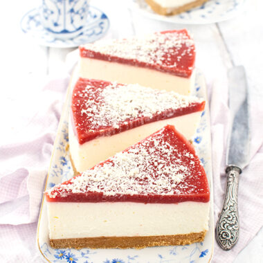 Drei Stücke Quark-Mascarpone-Torte auf einer einer weißen Porzellanplatte mit blauen Blümchen. Jedes Stück hat oben auf Erdbeermus und ist mit weißen Schokorapseln bestreut. Rechts neben dem Teller liegt ein Messer. Im Hintergrund ein Kuchenstück auf einem Teller und eine Tasse Kaffee