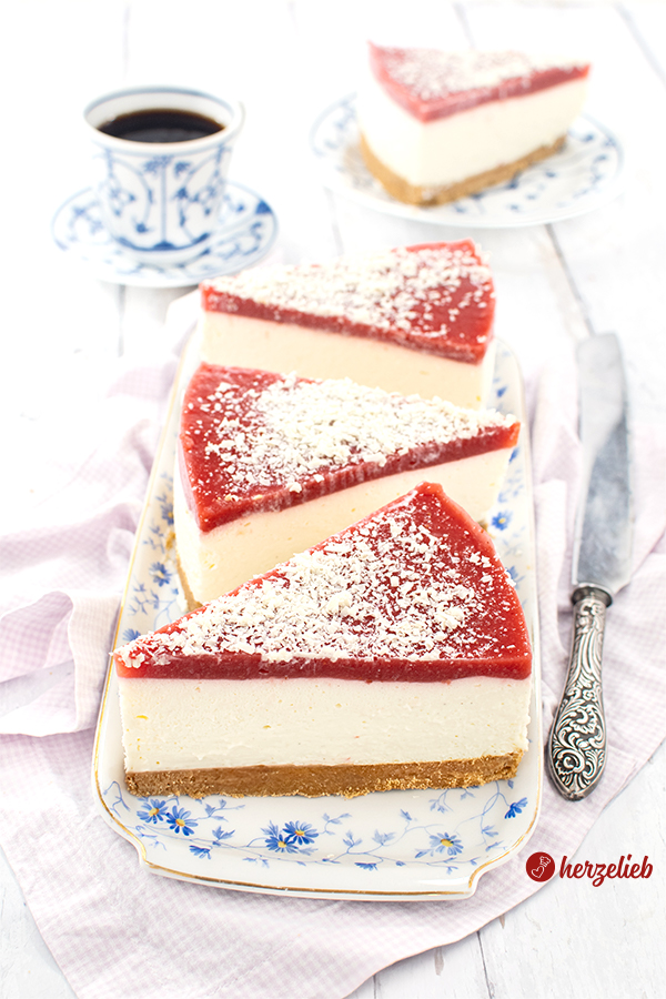 Drei Stücke Quark-Mascarpone-Torte auf einer einer weißen Porzellanplatte mit blauen Blümchen.  Jedes Stück hat oben auf Erdbeermus und ist mit weißen Schokorapseln bestreut. Rechts neben dem Teller liegt ein Messer. Im Hintergrund ein Kuchenstück auf einem Teller und eine Tasse Kaffee