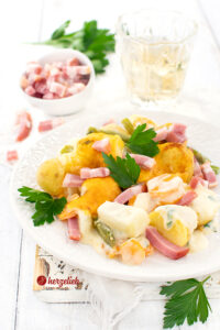 Ein Gemüseauflauf mit Kohlrabi, Karotten, Kartoffeln und Bohnen nach einem Schnüsch-Auflauf Rezept von herzelieb. Überbacken mit Käse und dekoriert mit Petersilie. Im Hintergrund ein Glas Saft, ein Petersilienbüschel und Bacon in einer kleinen, weißen Schale.