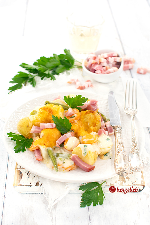 Ein Gemüseauflauf nach einem Schnüsch-Auflauf Rezept von herzelieb. Mit Käse überbacken und mit Kohlrabi, Karotten, Kartoffeln und Bohnen zubereitet. Überbacken mit Käse und dekoriert mit Petersilie. Rechts auf dem Tellerrand Silberbesteck. Im Hintergrund Petersilie und ein Schälchen Baconwürfel.