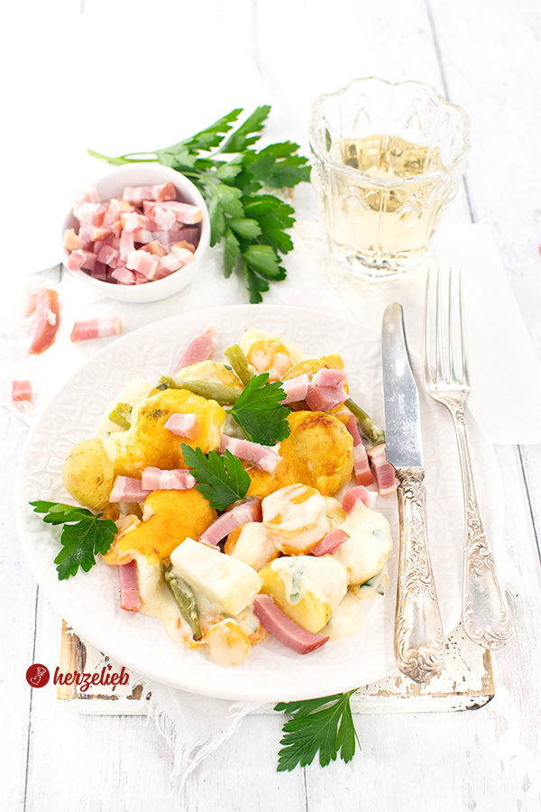 Ein weißer Teller mit einem Gemüseauflauf mit Bacon und Käse nach einem Schnüsch-Auflauf Rezept von herzelieb. Auf dem Tellerrand liegt Silberbesteck. Im Hintergrund ein Glas Saft, ein Büschel Petersilie und eine Schale mit Baconwürfeln. 