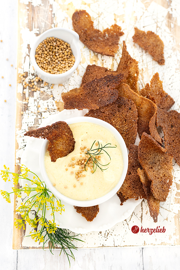 Ein Schälchen mit Honig-Senf-Dip, das dekoriert ist mit Dill und Senfkörnern im Vordergrund, um das selbst gemachte Schwarzbrotchips liegen. 