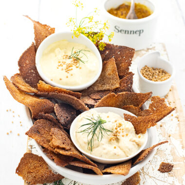 Eine Schale mit Schwarzbrotschips. In der Schale stehen zwischen den Brotchips zwei Schalen mit Honig-Senf-Dip. Im Hintergrund ein Schälchen mit Senfsaat und eines mit Senf.