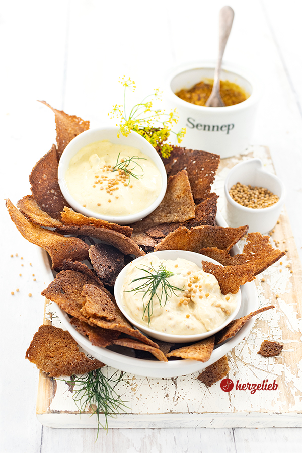 Eine Schale mit Schwarzbrotschips. In der Schale stehen zwischen den Brotchips zwei Schalen mit Honig-Senf-Dip. Im Hintergrund ein Schälchen mit Senfsaat und eines mit Senf.