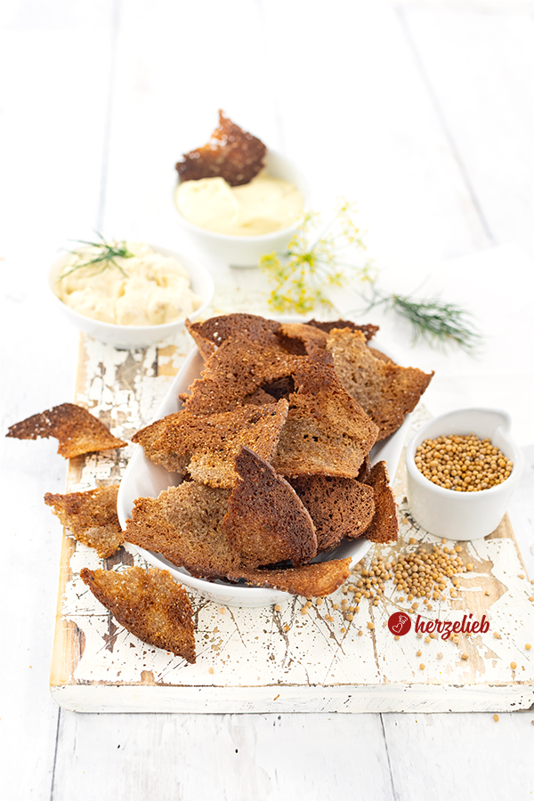 Eine längliche, weiße Porzellanschale mit Schwarzbrotchips. Rechts davon steh ein Schälchen mit Senfsaat. Im Hintergrund zwei Schalen mit Honig-Senf-Dip.