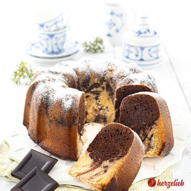 Ein angeschnittener, saftiger marmorierter Eierlikörkuchen mit echter Schokolade. Im Vordergrund zwei Schokoladenstücke. Ganz hinten blauweißes Kaffeegeschirr.