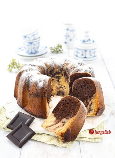 Ein angeschnittener, saftiger marmorierter Eierlikörkuchen mit echter Schokolade. Im Vordergrund zwei Schokoladenstücke. Ganz hinten blauweißes Kaffeegeschirr.