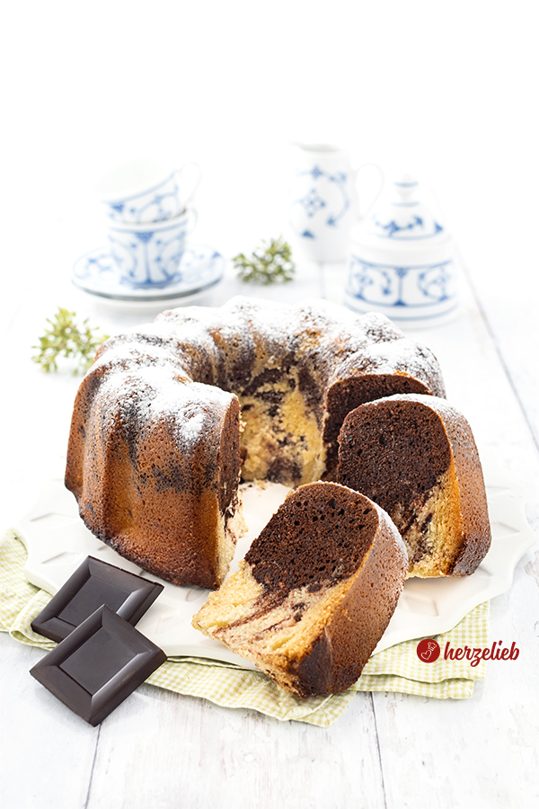 Ein angeschnittener, saftiger marmorierter Eierlikörkuchen mit echter Schokolade. Im Vordergrund zwei Schokoladenstücke. Ganz hinten blauweißes Kaffeegeschirr. 