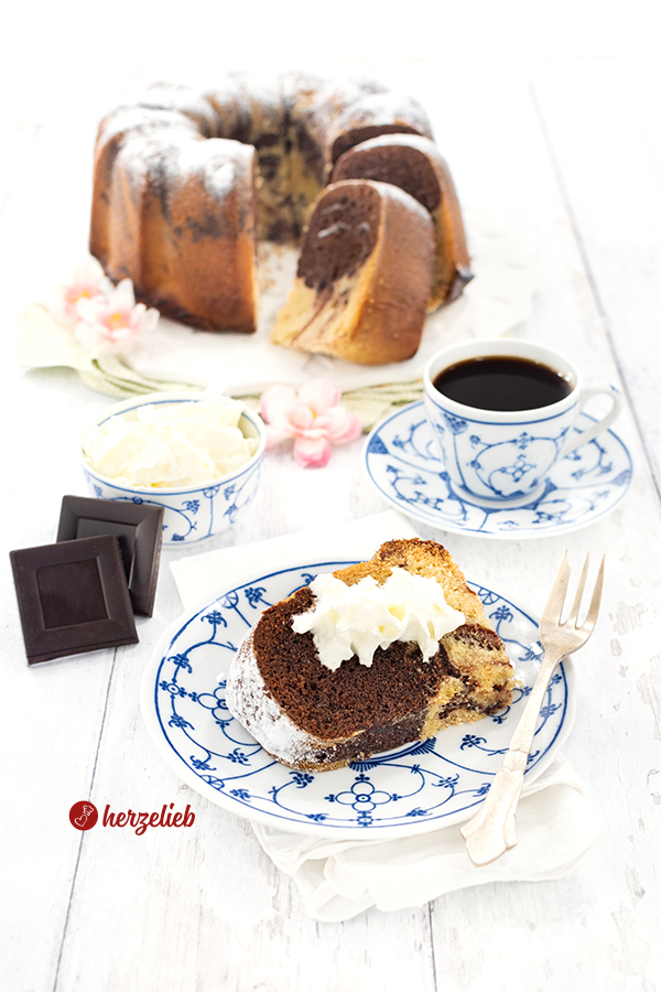 Ein Stück marmorierter Eierlikörkuchen nach einem Eierlikör-Marmorkuchen Rezept von herzelieb auf einem blauweißen Teller. Dahinter eine Tasse Kaffee und ein Schälchen Sahne. Ganz hinten der angeschnittene Kuchen.