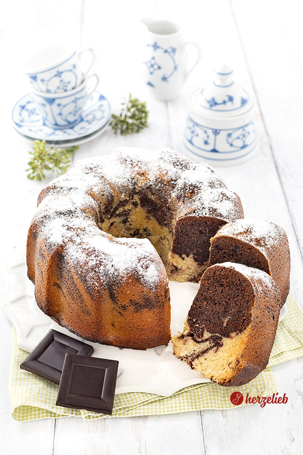 Ein angeschnittener marmorierter Eierlikörkuchen nach einem Eierlikör-Marmorkuchen Rezept von herzelieb. Vorne link liegen zwei Stücke Schokolade. Zwei Kuchenstücke sind nach vorne gezogen. Im Hintergrund steht blauweißes Kaffeegeschirr. 