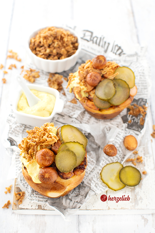 Zwei Hot Dog Muffins auf Zeitungspapier. Angerichtet mit dänischer Remoulade, dänischen Agurker und Röstzwiebeln. Zwischen dem Fingerfood steht ein Schälchen mit Remoulee und eines mti Röstzwiebeln. Verstreut liegen Wurststücke und Gurkenscheiben.