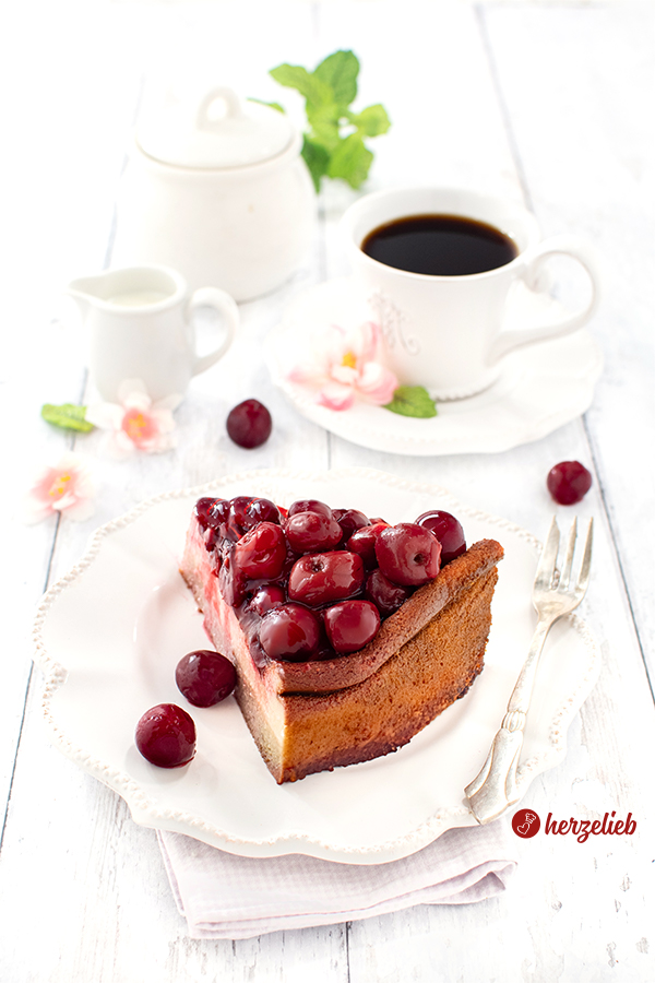 Käsekuchen mit Kartoffel-Mandelboden und Kirschen – angerichtet auf weißem Geschirr, im Hintergrund Kaffeegeschirr.