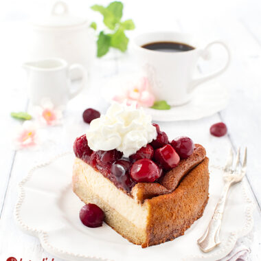 Ein Stück Cheescake nach einem Käsekuchen mit Kartoffel-Mandelboden von herzelieb. Angerichtet mit Sahne. Hinten dem Kuchenteller eine Tasse Kaffee, ein Zuckertopf, ein Milchkännchen, Blüten