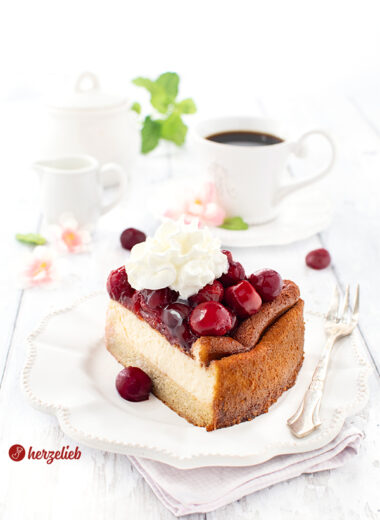 Ein Stück Cheescake nach einem Käsekuchen mit Kartoffel-Mandelboden von herzelieb. Angerichtet mit Sahne. Hinten dem Kuchenteller eine Tasse Kaffee, ein Zuckertopf, ein Milchkännchen, Blüten