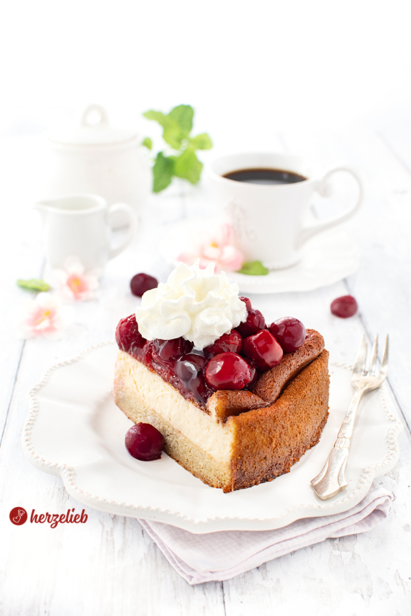 Ein Stück Cheescake nach einem Käsekuchen mit Kartoffel-Mandelboden von herzelieb. Angerichtet mit Sahne. Hinten dem Kuchenteller eine Tasse Kaffee, ein Zuckertopf, ein Milchkännchen, Blüten 