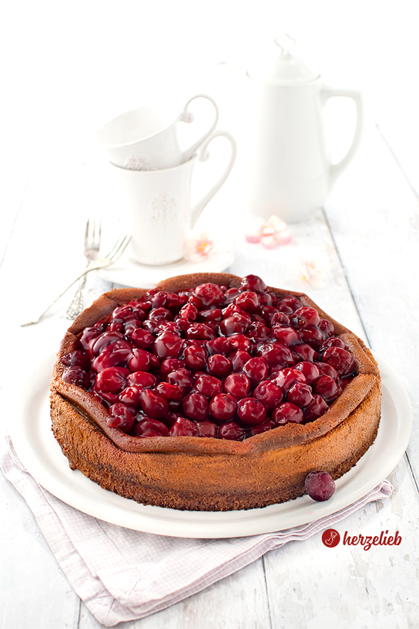 Ein ganzer Cheescake mit Kirschtopping nach einem Käsekuchen mit Kartoffel-Mandelboden Rezept. Der Kuchen ist goldbraun gebacken. Hinter dem Kuchenteller steht weißes Kaffeegeschirr