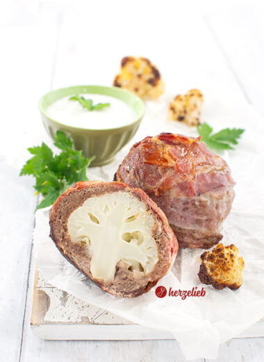 Zwei Blumenkohlbomben nach einem rezept von herzelieb. Das vordere Stück ist halbiert und man sieht im Inneren den Blumenkohl. Vorher rechts ein Stück gebratener Blumenkohl. Im Hintergrund ein grünes Schälchen mit Kräuterdip, davor liegt en Büschel Petersilie. Ganz hinten weitere geröstete Blumenkohlröschen