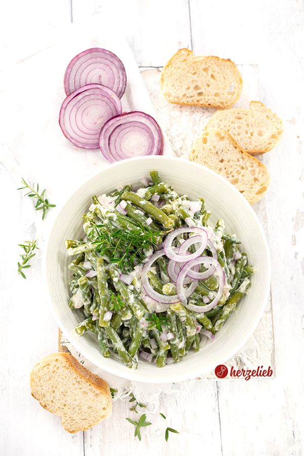 Sahne-Bohnensalat in einer weißen Schüssel, von oben fotografiert und angerichtet mit roten Zwiebelwürfeln und Zwiebelscheiben sowie Bohnenkraut. Im Hintergrund eine angschnittenen Zwiebel und einige Brotscheiben. Vorne ein weiteres Brot