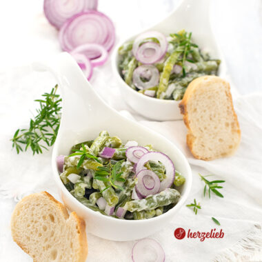 Zwei kleine, weiße Schüsselchen mit einem grünen Beilagensalat nach einem Sahne-Bohnensalat Rezept von herzelieb. Dekoriert mit roten Zwiebelringen, Zwiebelwürfeln nd Bohnenkraut. Angerichtet mit Brot.