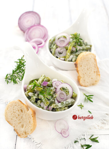 Zwei kleine, weiße Schüsselchen mit einem grünen Beilagensalat nach einem Sahne-Bohnensalat Rezept von herzelieb. Dekoriert mit roten Zwiebelringen, Zwiebelwürfeln nd Bohnenkraut. Angerichtet mit Brot.