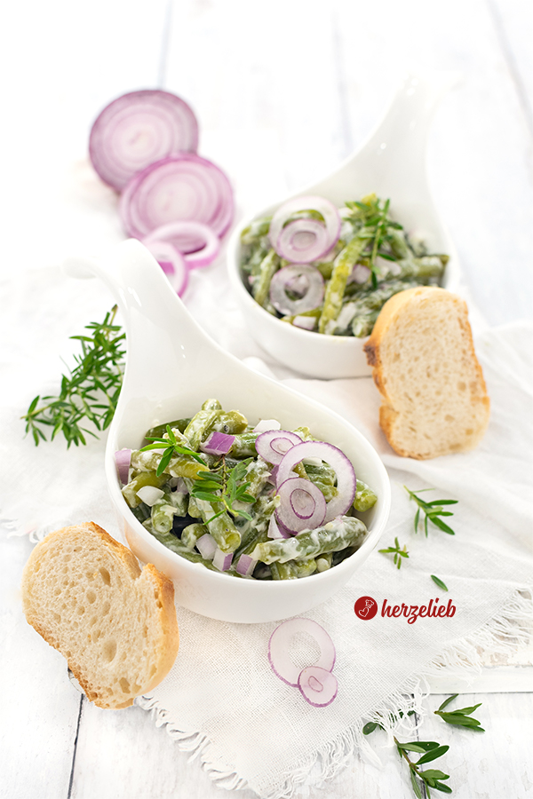 Zwei kleine, weiße Schüsselchen mit einem grünen Beilagensalat nach einem Sahne-Bohnensalat Rezept von herzelieb. Dekoriert mit roten Zwiebelringen, Zwiebelwürfeln nd Bohnenkraut. Angerichtet mit Brot.