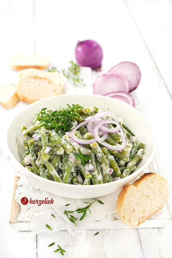 Eine Schüssel mit einem grünem Beilagensalat nach Omas Sahne-Bohnensalat Rezept von herzelieb. Angerichtet mit roten Zwiebelnwürfeln und -scheiben und Bohnenkraut. Vorne rechts ein Stück Brot. Hinten eine aufgeschnittene und eine ganze, rote Zwiebel, Kräuter und Brotscheiben.