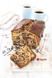 Ein angeschnittener Kastenkuchen nach einem Ameisenkuchen Rezept von herzelieb auf einem weißen Holzbrett. Vor dem schräg stehendem Rührkuchen liegt ein halbes Stück davon. Es liegen Schokostreusel daneben. Rechts vom Kuchen liegt eine Kuchengabel. Im Hintergrund zwei Becher Kaffee und ein Geschirrtuch.