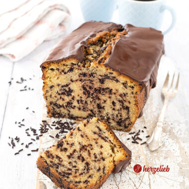 Ein angeschnittener Kastenkuchen nach einem Ameisenkuchen Rezept von herzelieb auf einem weißen Holzbrett. Vor dem schräg stehendem Rührkuchen liegt ein halbes Stück davon. Es liegen Schokostreusel daneben. Rechts vom Kuchen liegt eine Kuchengabel. Im Hintergrund zwei Becher Kaffee und ein Geschirrtuch.