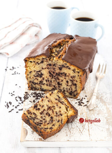 Ein angeschnittener Kastenkuchen nach einem Ameisenkuchen Rezept von herzelieb auf einem weißen Holzbrett. Vor dem schräg stehendem Rührkuchen liegt ein halbes Stück davon. Es liegen Schokostreusel daneben. Rechts vom Kuchen liegt eine Kuchengabel. Im Hintergrund zwei Becher Kaffee und ein Geschirrtuch.