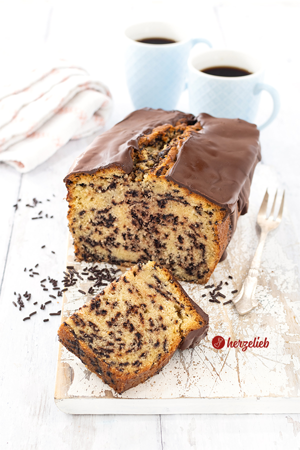 Ein angeschnittener Kastenkuchen nach einem Ameisenkuchen Rezept von herzelieb auf einem weißen Holzbrett. Vor dem schräg stehendem Rührkuchen liegt ein halbes Stück davon. Es liegen Schokostreusel daneben. Rechts vom Kuchen liegt eine Kuchengabel. Im Hintergrund zwei Becher Kaffee und ein Geschirrtuch.