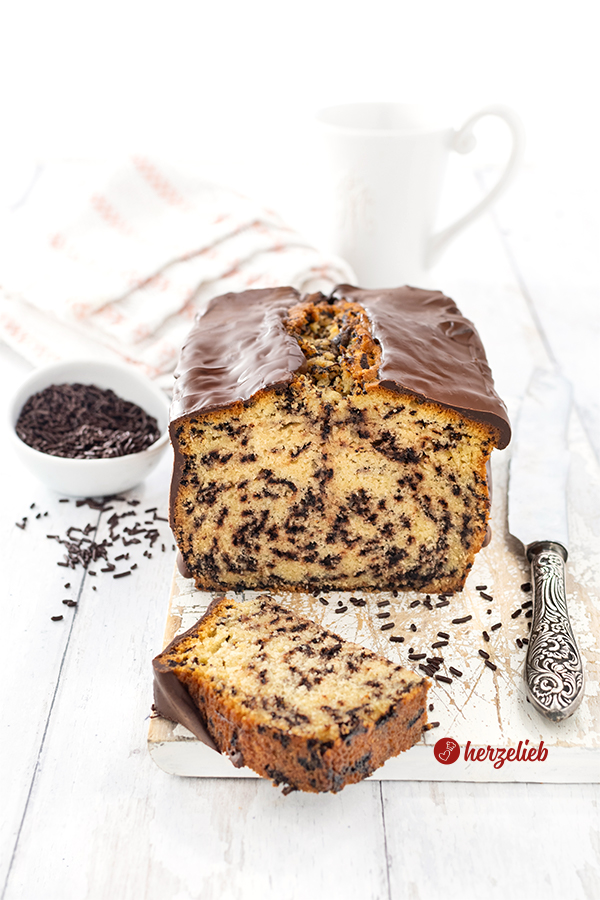 Ein angeschnittener Rührkuchen mit Schokoladenstreuseln nach einem Ameisenkuchen Rezept von herzelieb. Der angeschnittene Kuchen mit Schokoladenglasur liegt auf einem weißen Holzbrett . Rechts daneben liegt ein Messer. Im Hintergrund eine Kaffeetasse und ein Geschirrtuch. Links ein Schälchen mit Schokostreuseln.