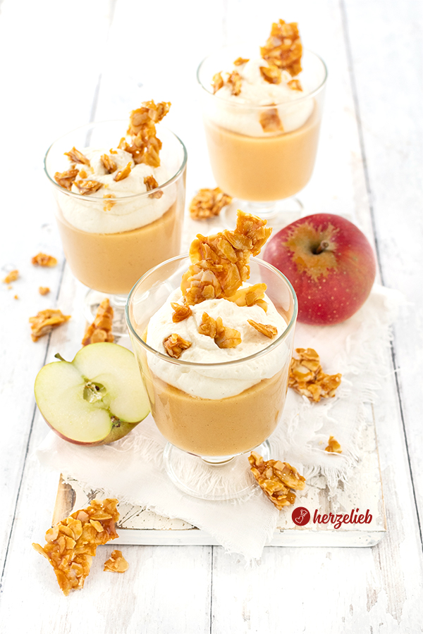 Drei Gläser mit einem Apfeldessert im Glas. Mit einem Topping aus Schmandsahne und Mandelkrokant. Zwischen den Gläsern liegt wieterer Krokant, ein Apfel und weiter vorne ein halbierter Apfel.