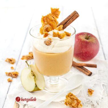 Ein Glas mit Apfelcreme-Dessert, angerichtet in einem Wasserglas Sahne-Schmand obendrauf. Dekoriert mit Mandelkrokant und einer Zimtstange.