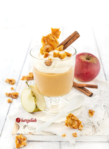 Ein Glas mit Apfelcreme-Dessert, angerichtet in einem Wasserglas Sahne-Schmand obendrauf. Dekoriert mit Mandelkrokant und einer Zimtstange.