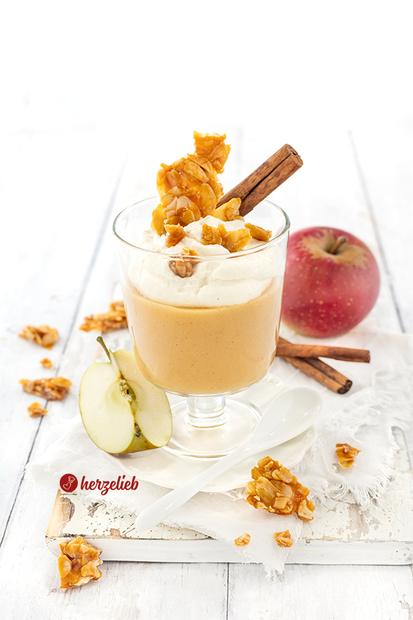 Ein Glas mit Apfelcreme-Dessert, angerichtet in einem Wasserglas  Sahne-Schmand obendrauf. Dekoriert mit Mandelkrokant und einer Zimtstange. 