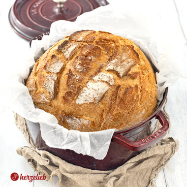 Ein Dinkelbrot nach einem Dinkel-Joghurt-Brot Rezept von herzelieb. Das Brot liegt in einem roten Topf, der auf einen braunen Tuch liegt. Im Hintergrund liegt der Deckel