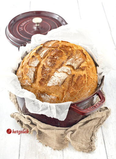 Ein Dinkelbrot nach einem Dinkel-Joghurt-Brot Rezept von herzelieb. Das Brot liegt in einem roten Topf, der auf einen braunen Tuch liegt. Im Hintergrund liegt der Deckel