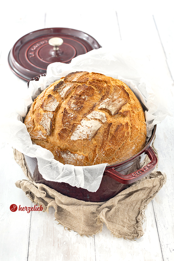 Ein Dinkelbrot nach einem Dinkel-Joghurt-Brot Rezept von herzelieb. Das Brot liegt in einem roten Topf, der auf einen braunen Tuch liegt. Im Hintergrund liegt der Deckel