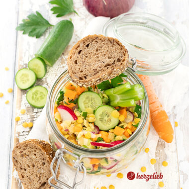 Ein Bügelglas oder WEckglas mit einem bunten Salat nach einem gelber Linsensalat Rezept von herzelieb. Zubereitet mit Karotten, Radieschen, Linsen, Petersilie, Zwiebel und Sellerie. Im Hintergrund eine rote Zwiebel, einige Petersilienblättchen, eine angeschnittene Gurke Vorne links liegen zwei Brotscheiben