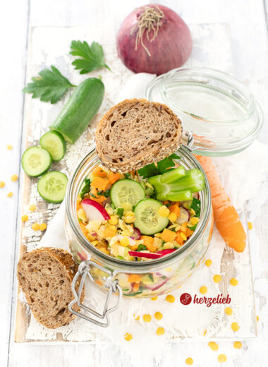 Ein Bügelglas oder WEckglas mit einem bunten Salat nach einem gelber Linsensalat Rezept von herzelieb. Zubereitet mit Karotten, Radieschen, Linsen, Petersilie, Zwiebel und Sellerie. Im Hintergrund eine rote Zwiebel, einige Petersilienblättchen, eine angeschnittene Gurke Vorne links liegen zwei Brotscheiben