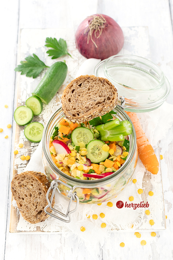 Ein Bügelglas oder WEckglas mit einem bunten Salat nach einem gelber Linsensalat Rezept von herzelieb. Zubereitet mit Karotten, Radieschen, Linsen, Petersilie, Zwiebel und Sellerie. Im Hintergrund eine rote Zwiebel, einige Petersilienblättchen, eine angeschnittene Gurke Vorne links liegen zwei Brotscheiben