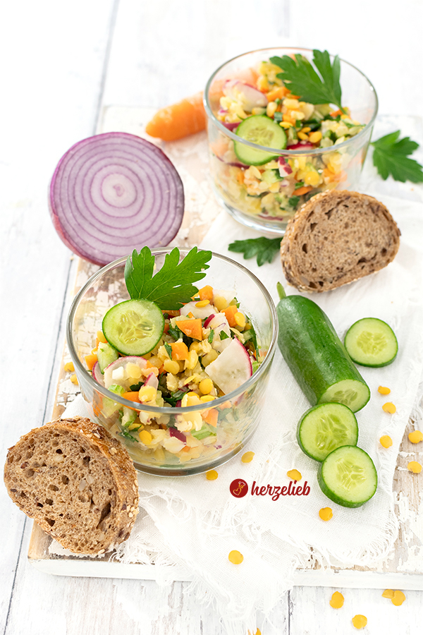 Zwei Gläser mit einem bunten Gemüsesalat mit Linsen nach einem gelber Linsensalat Rezept von herzelieb. Rechts im Bild eine angeschnittene Gurke, hinter dem vorderen Glas eine halbe rote Zwiebel. Weiter hinten eine Karotte und eine Scheibe Brot. Vorne links sind zwei Brotscheiben zu sehen. 