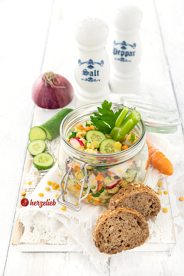 Ein bunter Salat mit viel Gemüse nach einem Gelber Linsensalat Rezept von herzelieb. Angerichtet in einem Bügelglas, dekoriert mit Gurkenscheiben und Petersilie. Im Hintergrund stehen ein Salz- und ein Pfefferstreuer, eine halbe rote Zwiebel und eine angeschnittene Gurke. Vorne rechts liegen zwei Brotscheiben. 