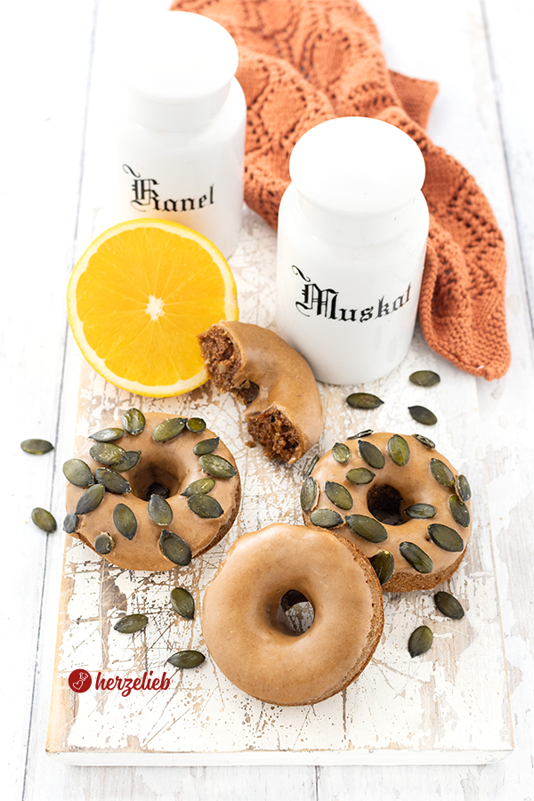 Drei verschiedene kleine Kürbiskuchen nach einem Kürbis-Donuts Rezept von herzelieb. Alle Donuts haben einen Zimtguss, zwei davon sind mit Kürbiskernen bestreut. Im Hintergrund ein halber Minikuchen. Dahinter eine halbe Orange, zwei Gewürzgefäße und und braunes Tuch.