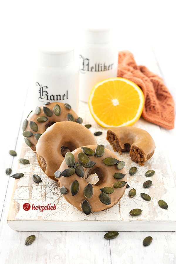 Drei kleine Kürbiskuchen mit einer Zimtglasur nach einem Kürbis-Donuts Rezept von herzelieb. Zwei der Donuts sind mit Kürbiskernen bestreut.  Im Hintergrund liegt ein halber Donut, eine halbe Orange und zwei Gewürzgefäße aus Porzellan.